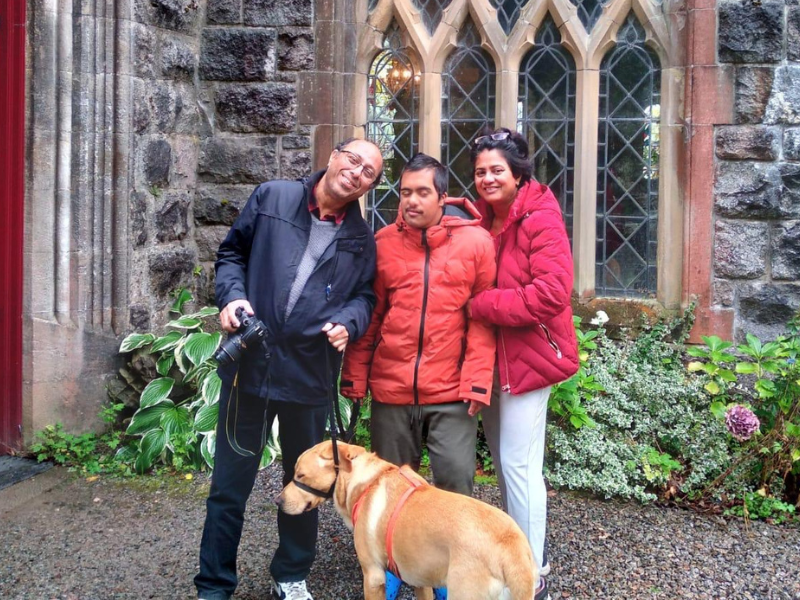 Aaryan, mum Pronita-and dad Subhayu stand outside a historic building with their dog. They are wearing warm jackets and smiling at the camera.