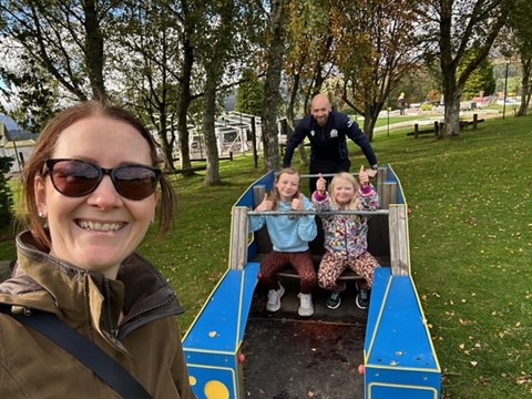 A family of four outside in the sunshine using play equipment